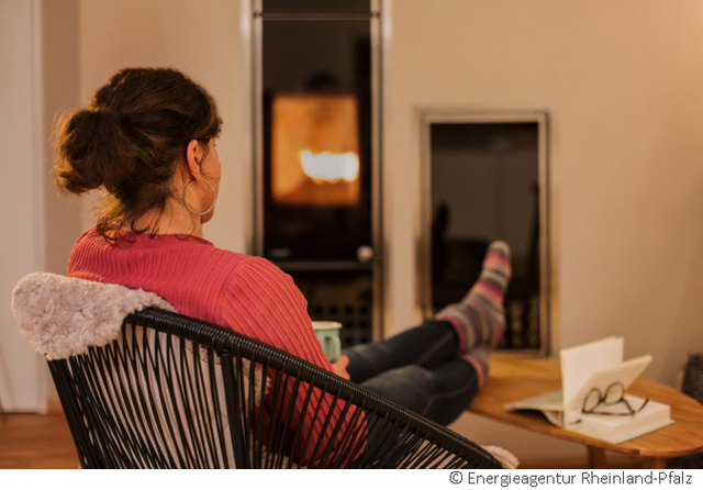 Eine Person sitzt im Wohnzimmer vor einem Pelletsofen mit loderndem Feuer.