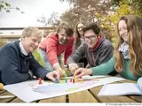 Auf einem Tisch liegt eine Karte von Rheinland-Pfalz. Fünf Menschen sitzen um den Tisch und platzieren auf der Karte bunte Kegel in menschenähnlicher Form.