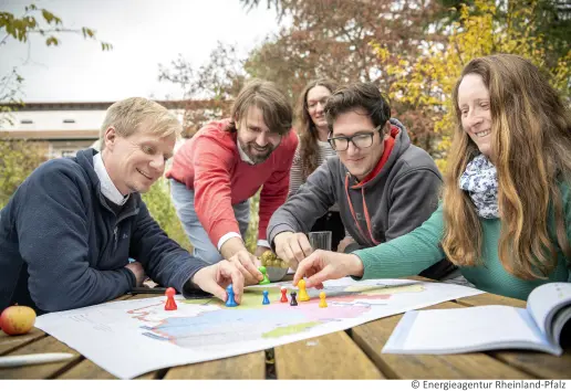 Auf einem Tisch liegt eine Karte von Rheinland-Pfalz. Fünf Menschen sitzen um den Tisch und platzieren auf der Karte bunte Kegel in menschenähnlicher Form.