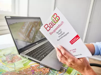 Laptop steht auf Bebauungsplan, Hände halten Bauggesetzbuch