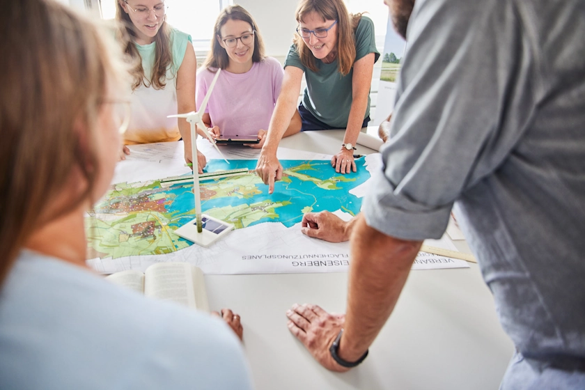 Mehrere Menschen sind um einen Tisch versammelt und sehen auf einen Flächennutzungsplan. Darauf steht auch das Modell eines Windrades.