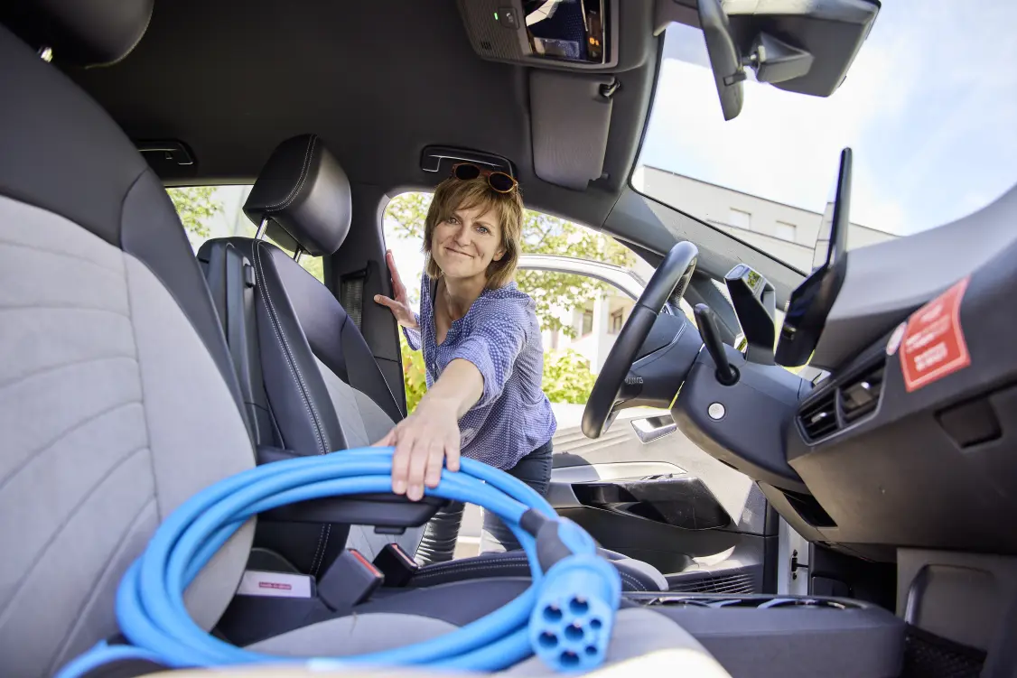 Eine Person blickt auf der Fahrerseite ins Auto und holt vom Beifahrersitz des Autos das Ladekabel.