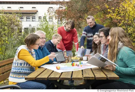 Auf einem Tisch liegt eine Karte von Rheinland-Pfalz. Mehrere Menschen sitzen um den Tisch und besprechen etwas. Eine Person zeigt auf die Abbildung auf einem tablet.