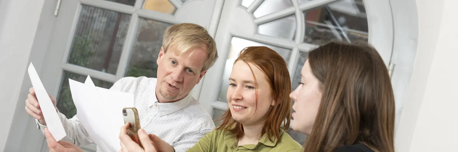 Drei Personen besprechen etwas. Sie blicken auf zwei Papierblätter und ein Handy.